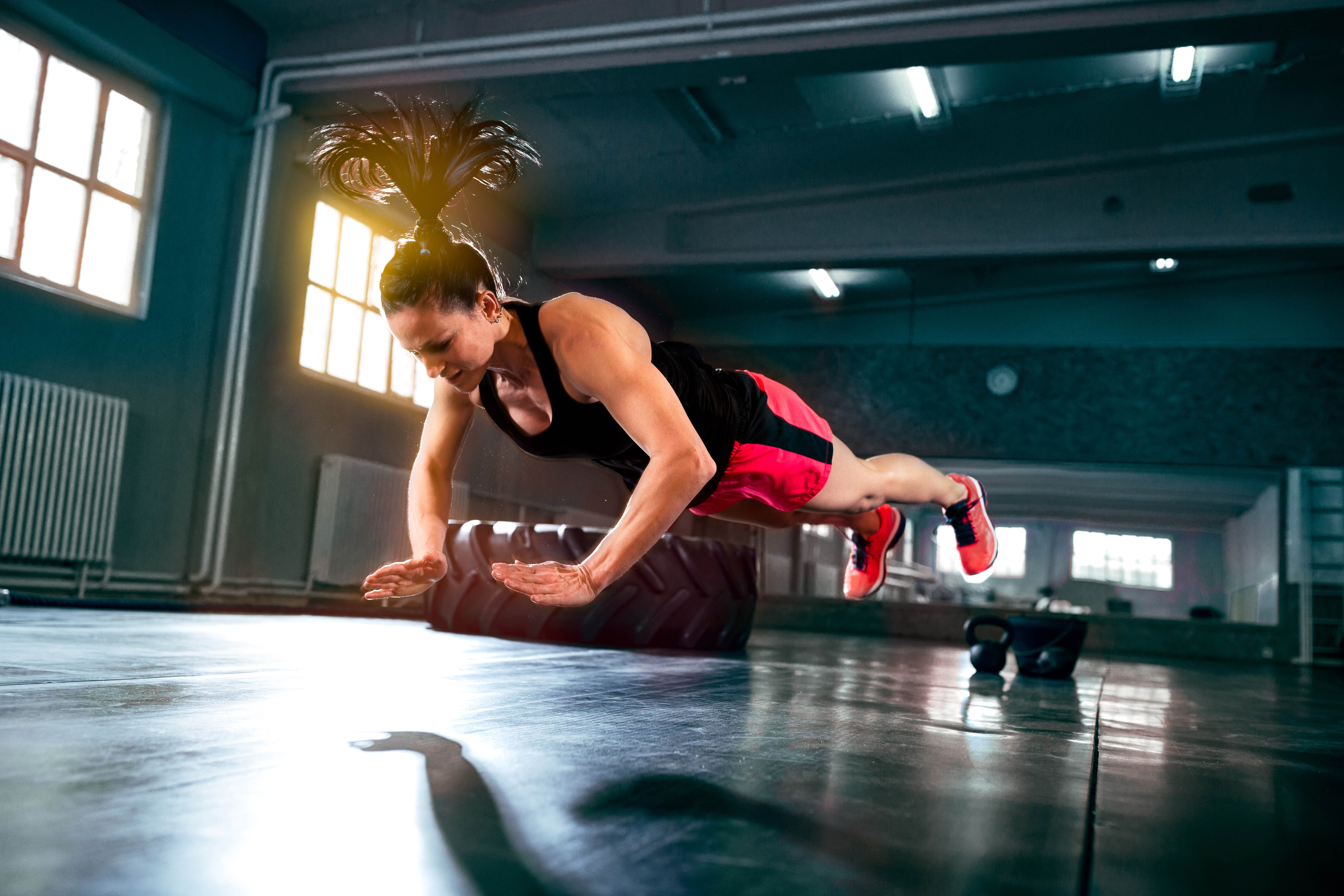 Young, fit woman mid-flight during a clapping push-up. Background is a gym, light streaming in through the window.